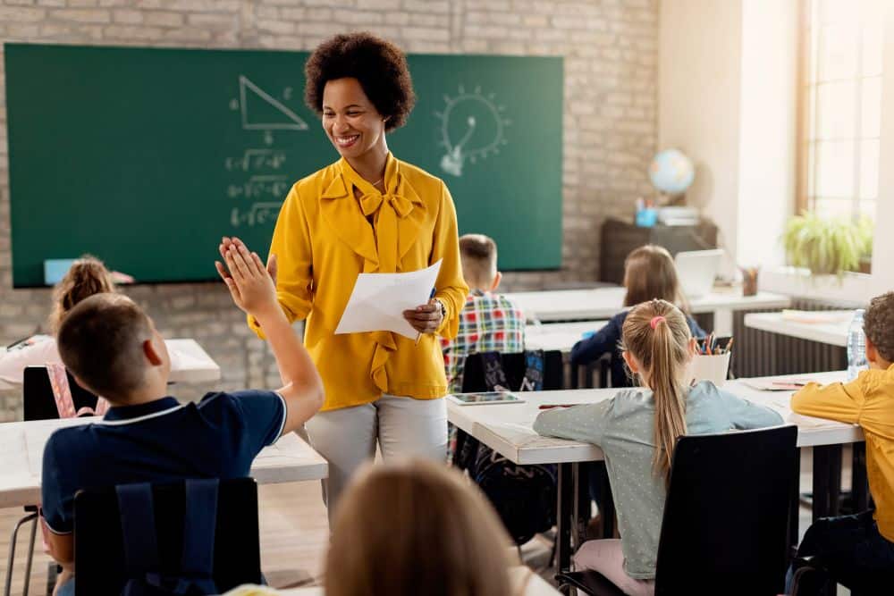 Uma professora negra sorridente, vestindo blusa amarela, cumprimenta um aluno com um "high five" em uma sala de aula iluminada. Ao fundo, um quadro verde com desenhos de geometria e outros alunos sentados em suas carteiras.