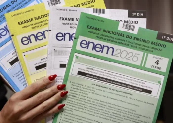 Mão feminina segura cadernos coloridos do Enem 2025, nas cores azul, amarelo, branco e verde.