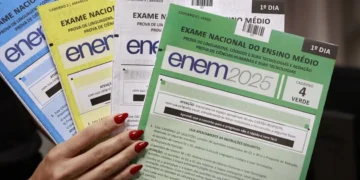 Mão feminina segura cadernos coloridos do Enem 2025, nas cores azul, amarelo, branco e verde.