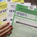 Mão feminina segura cadernos coloridos do Enem 2025, nas cores azul, amarelo, branco e verde.