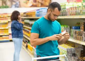 Homem verificando rótulo de produto em supermercado, com mulher ao fundo, refletindo mudanças na operação de supermercados aos domingos.