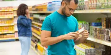 Homem verificando rótulo de produto em supermercado, com mulher ao fundo, refletindo mudanças na operação de supermercados aos domingos.