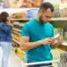 Homem verificando rótulo de produto em supermercado, com mulher ao fundo, refletindo mudanças na operação de supermercados aos domingos.