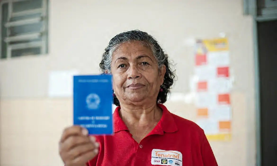 Valorização do Trabalho Doméstico em 2026: Direitos e Conquistas Mulher com crachá da Fenatrad segura carteira de trabalho azul em destaque.