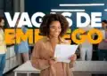 Mulher negra sorridente com cabelos cacheados, vestindo camisa bege, segurando papéis e uma caneta em um escritório. Sobre a imagem, o texto em destaque diz "VAGAS DE EMPREGO" em cores branco e amarelo. Ao fundo, outras pessoas desfocadas trabalham no ambiente.