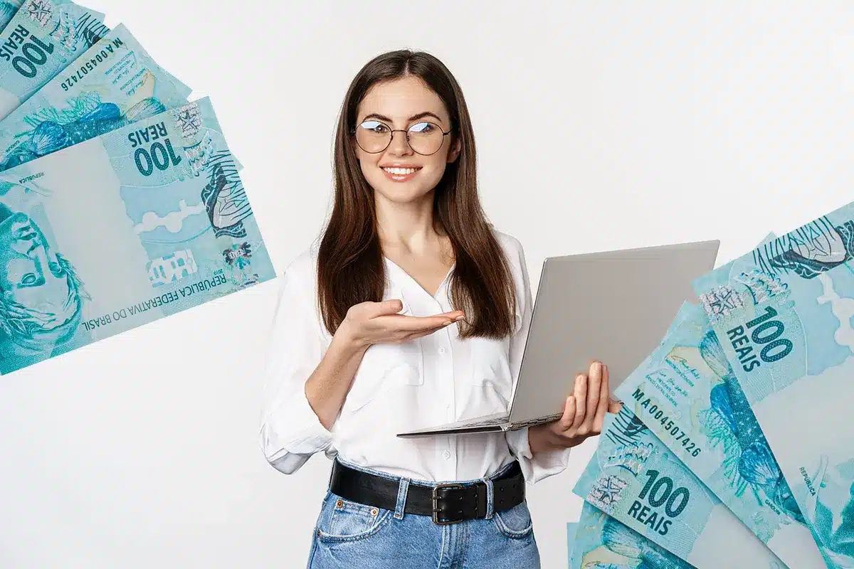Jovem com notebook destaca oportunidades de renda digital em 2026 Jovem sorrindo segura notebook e aponta para notas de cem reais brasileiras ao redor