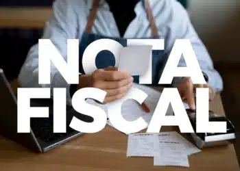 Pessoa segurando nota fiscal diante de notebook e máquina registradora em mesa de madeira