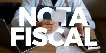 Pessoa segurando nota fiscal diante de notebook e máquina registradora em mesa de madeira