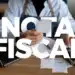 Pessoa segurando nota fiscal diante de notebook e máquina registradora em mesa de madeira