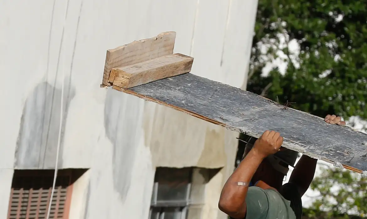 Trabalhador carregando tábua em obra de construção civil Homem segura tábua de madeira sobre os ombros em frente a parede de prédio