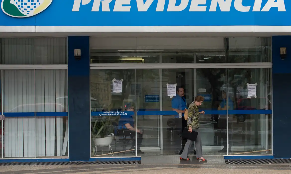 Fachada de agência do INSS durante horário de atendimento Mulher idosa sai por porta de vidro da agência do INSS enquanto funcionários atendem no interior