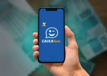 Mão segura celular exibindo tela inicial do app Caixa Tem sobre notas de cem reais desfocadas