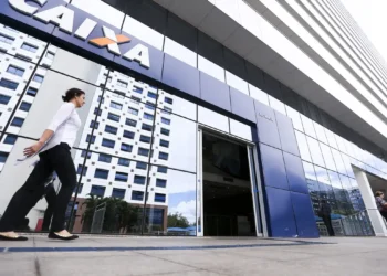 Mulher caminha em frente à entrada de agência da Caixa com prédio espelhado ao fundo