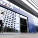 Mulher caminha em frente à entrada de agência da Caixa com prédio espelhado ao fundo