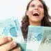 Mulher jovem sorrindo exibe várias notas de cem reais com destaque para o dinheiro nas mãos.
