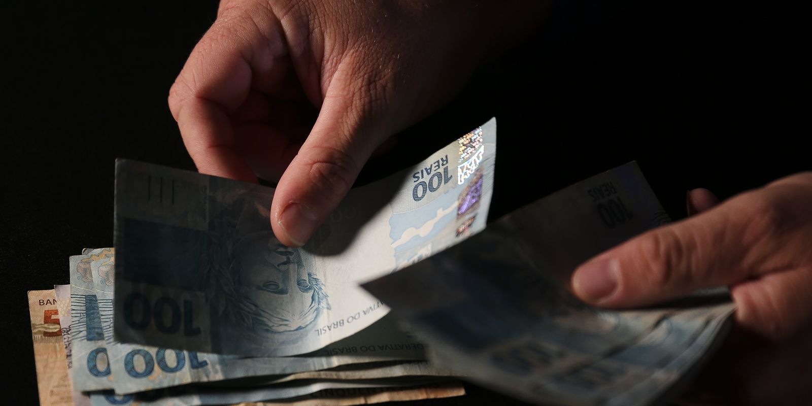 Mãos humanas contando notas de cem reais sob fundo escuro, indicando recebimento de valores