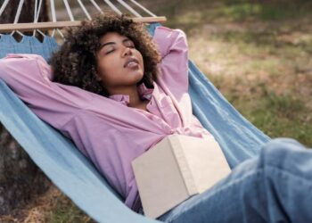 Imagem mostra uma mulher relaxando em uma rede ao ar livre enquanto lê um livro, representando descanso e aproveitamento do tempo livre durante feriados prolongados no mês de abril.