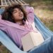 Imagem mostra uma mulher relaxando em uma rede ao ar livre enquanto lê um livro, representando descanso e aproveitamento do tempo livre durante feriados prolongados no mês de abril.