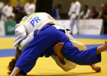 Dois atletas de judô competem no tatame, um aplicando golpe de queda com uniforme azul.
