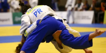 Dois atletas de judô competem no tatame, um aplicando golpe de queda com uniforme azul.