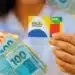 Mão segura cartão do Bolsa Família ao lado de várias cédulas de reais coloridas
