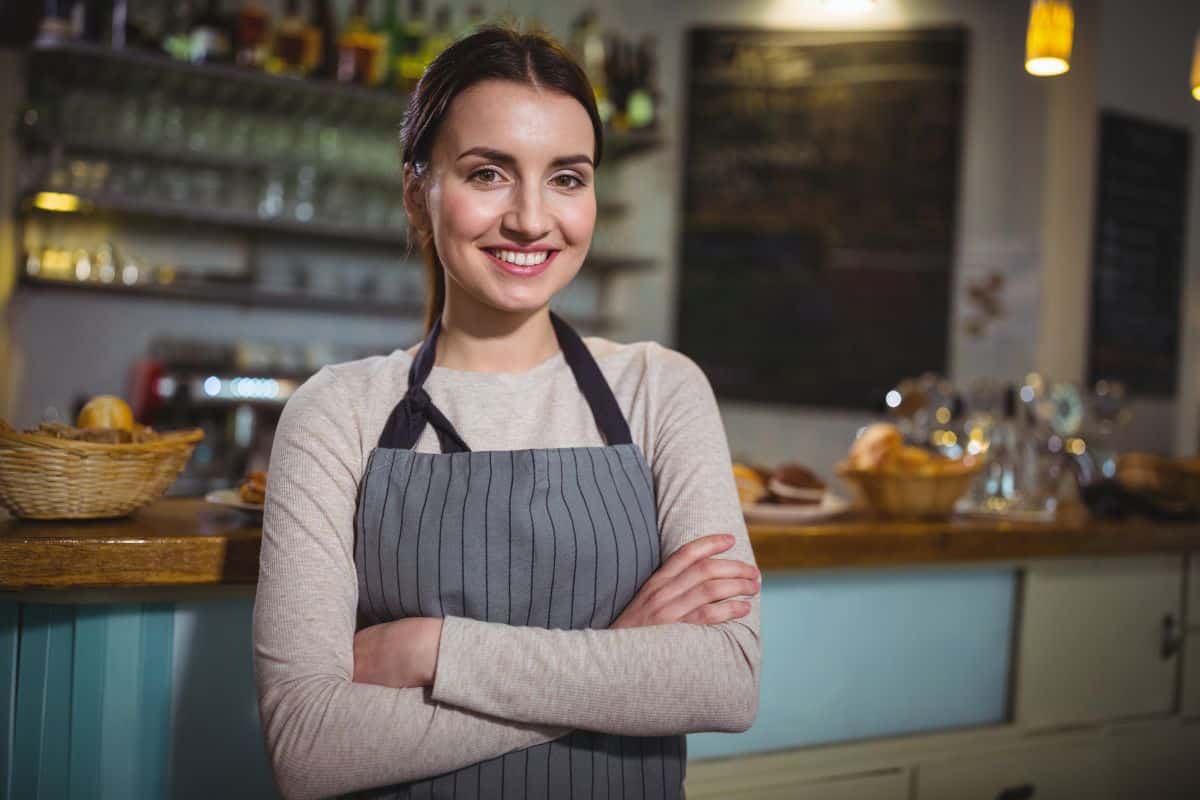 Mulher sorrindo de avental cinza com braços cruzados em frente ao balcão de uma cafeteria.