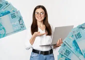 Jovem de óculos segura notebook e aponta para notas de R$100 ao redor.