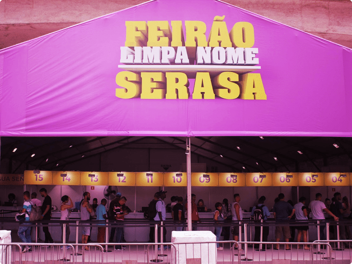 Entrada de uma grande tenda cor-de-rosa onde está montado o Feirão Limpa Nome Serasa. O letreiro na fachada é grande, amarelo e branco. Abaixo, há uma fila de pessoas em pé aguardando atendimento diante de vários guichês numerados e iluminados.