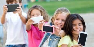Grupo de crianças sorrindo enquanto tiram selfies com celulares ao ar livre.