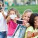Grupo de crianças sorrindo enquanto tiram selfies com celulares ao ar livre.