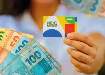 Mão segurando o cartão do Programa Bolsa Família ao lado de notas de R$ 100, R$ 50 e R$ 20, representando o pagamento do benefício em março de 2026