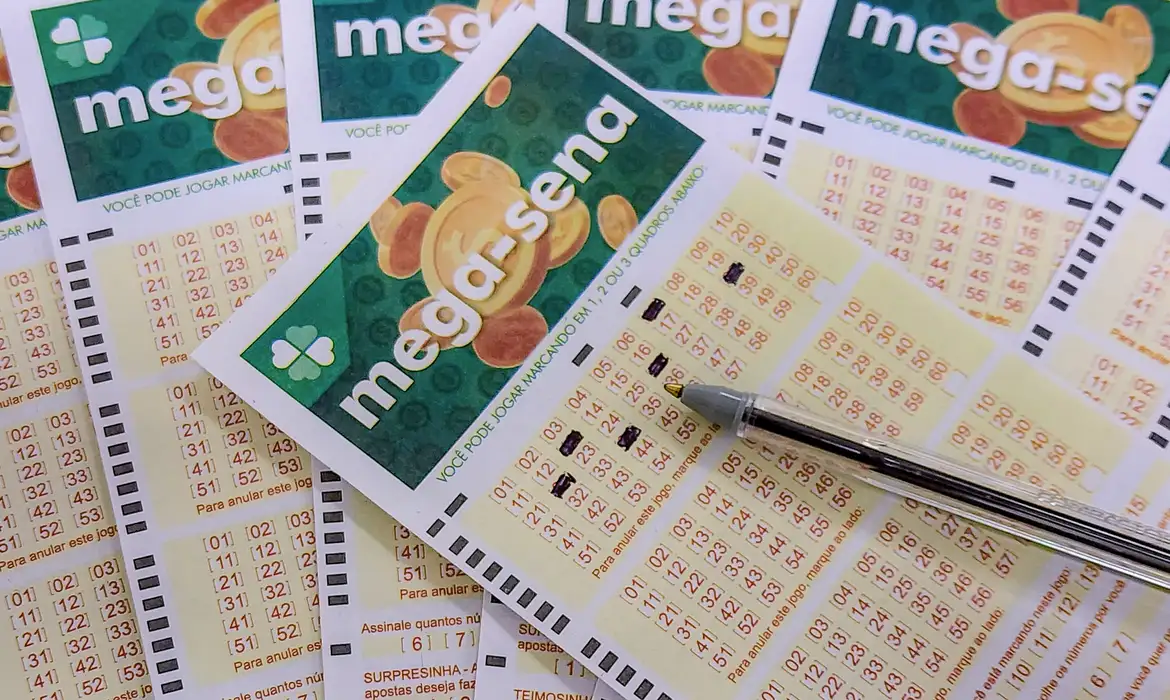 passo a passo como preencher bilhete mega sena Close em primeiro plano de uma caneta esferográfica preenchendo os quadrados de números em um volante físico da Mega-Sena, com outros bilhetes sobrepostos ao fundo.