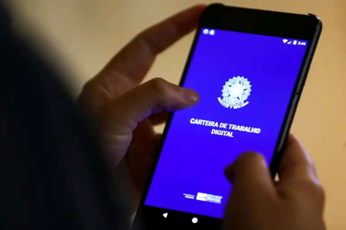 Mãos segurando um smartphone que exibe a tela azul do aplicativo Carteira de Trabalho Digital, com o logotipo do Governo Federal.