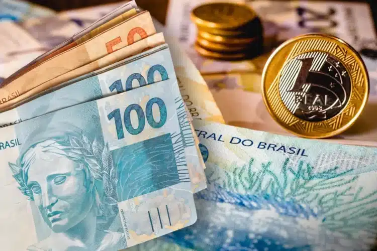 Imposto de Renda 2026 Notas de 100 e 50 reais junto a moedas de 1 real empilhadas sobre mesa