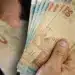 Mão segurando cédulas de cinquenta reais em contagem detalhada