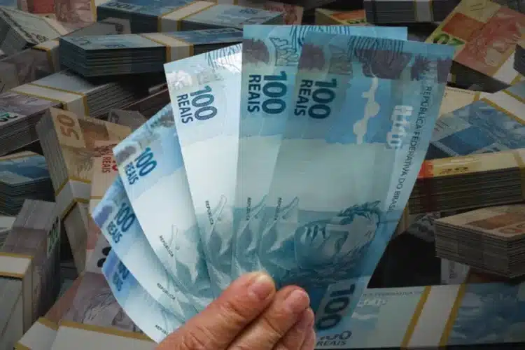 PIS/Pasep 2026 Mão segurando várias notas de R$ 100 reais com maços de dinheiro ao fundo