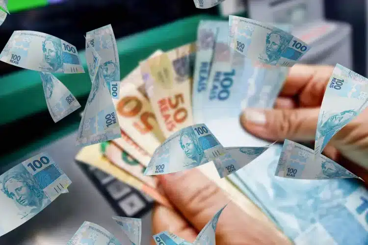 Isenção Imposto de Renda 2026 Mãos contam notas de 100 e 50 reais com cédulas voando em frente a caixa eletrônico