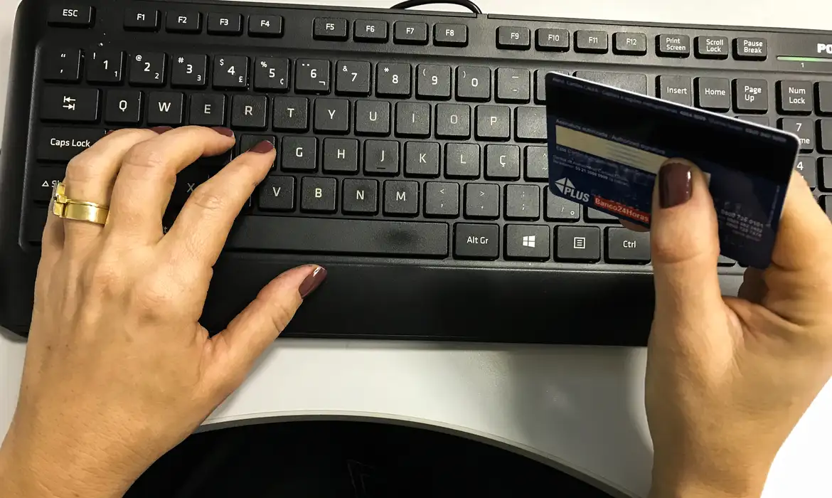 Renegociação de dívidas online no Mutirão Febraban 2026: acesso pelo site ou app Mãos femininas digitando em teclado de computador enquanto seguram cartão bancário, representando a negociação de dívidas online no Mutirão Febraban 2026 pelos canais digitais dos bancos