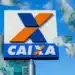 Placa da Caixa Econômica Federal com notas de R$ 100 voando ao redor em céu azul