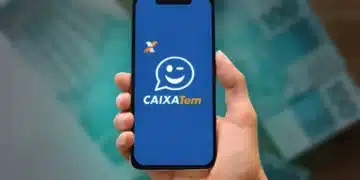 Mão segurando smartphone com tela de abertura do aplicativo Caixa Tem e cédulas desfocadas ao fundo — aplicativo usado para acessar o pagamento do Pé-de-Meia 2026