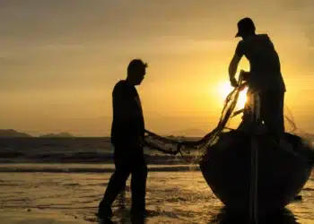 ois trabalhadores puxando rede de pesca na praia com barco ao entardecer