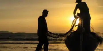 ois trabalhadores puxando rede de pesca na praia com barco ao entardecer
