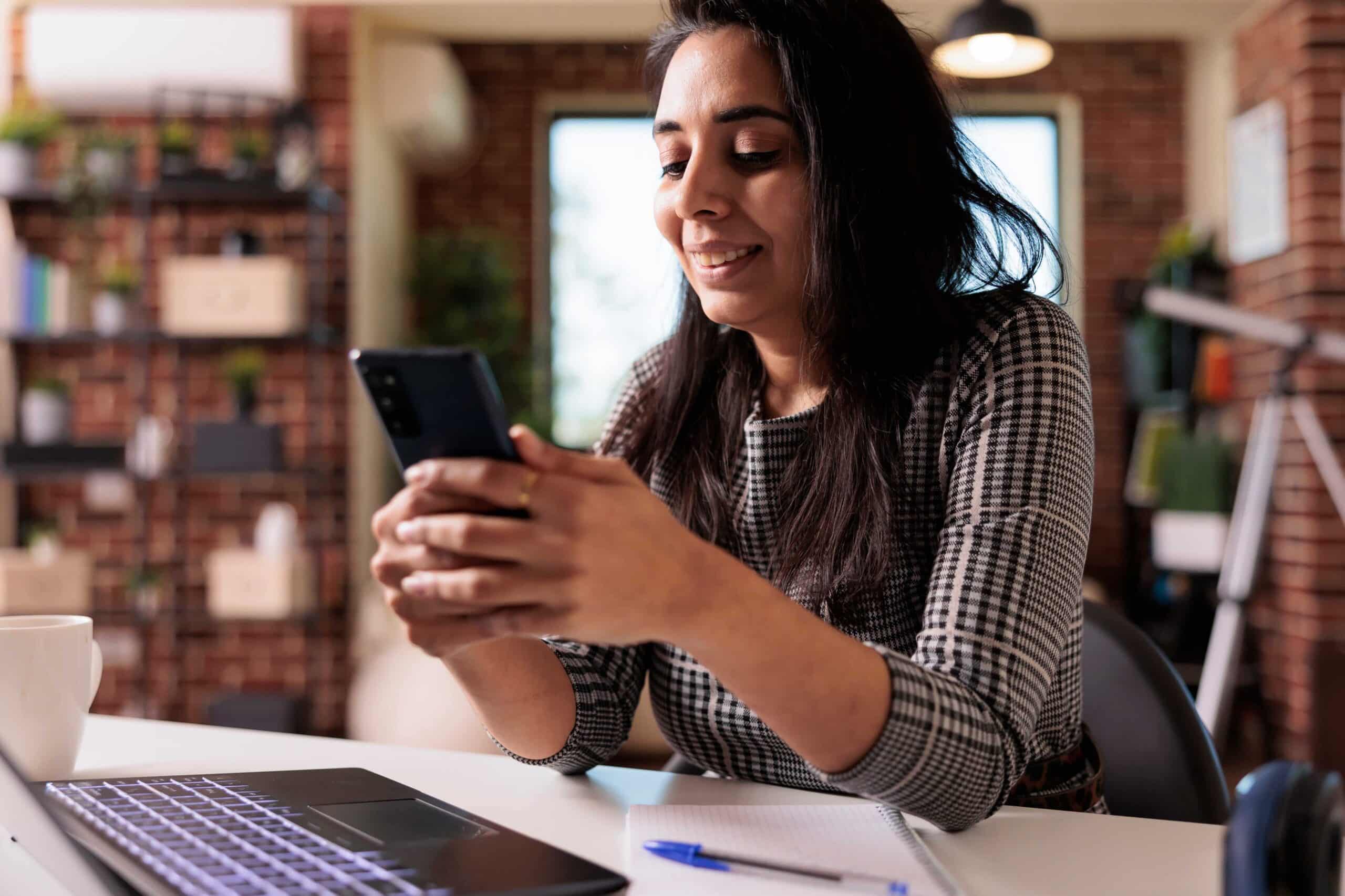 Mulher sorridente acessando informações pelo celular ao lado de notebook e caderno
