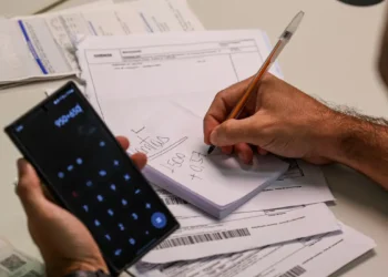 Homem segurando celular com calculadora aberta enquanto anota valores em bloco de papel sobre mesa com boletos e documentos financeiros, representando o planejamento para renegociação de dívidas no Mutirão Febraban 2026