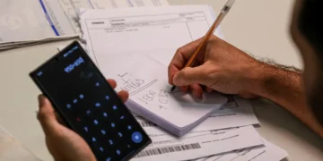 Homem segurando celular com calculadora aberta enquanto anota valores em bloco de papel sobre mesa com boletos e documentos financeiros, representando o planejamento para renegociação de dívidas no Mutirão Febraban 2026