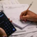 Homem segurando celular com calculadora aberta enquanto anota valores em bloco de papel sobre mesa com boletos e documentos financeiros, representando o planejamento para renegociação de dívidas no Mutirão Febraban 2026