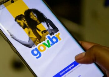 Close-up de uma mão segurando um smartphone que exibe a tela inicial de login do portal gov.br, mostrando o logotipo colorido sobre um fundo branco e amarelo com a imagem de um casal ao fundo.