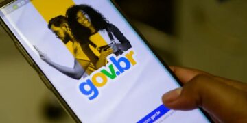 Close-up de uma mão segurando um smartphone que exibe a tela inicial de login do portal gov.br, mostrando o logotipo colorido sobre um fundo branco e amarelo com a imagem de um casal ao fundo.