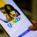 Close-up de uma mão segurando um smartphone que exibe a tela inicial de login do portal gov.br, mostrando o logotipo colorido sobre um fundo branco e amarelo com a imagem de um casal ao fundo.