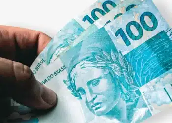 Close detalhado de uma mão segurando um maço com diversas cédulas azuis de 100 reais, com o texto "REPÚBLICA FEDERATIVA DO BRASIL" e o número "100" em destaque.